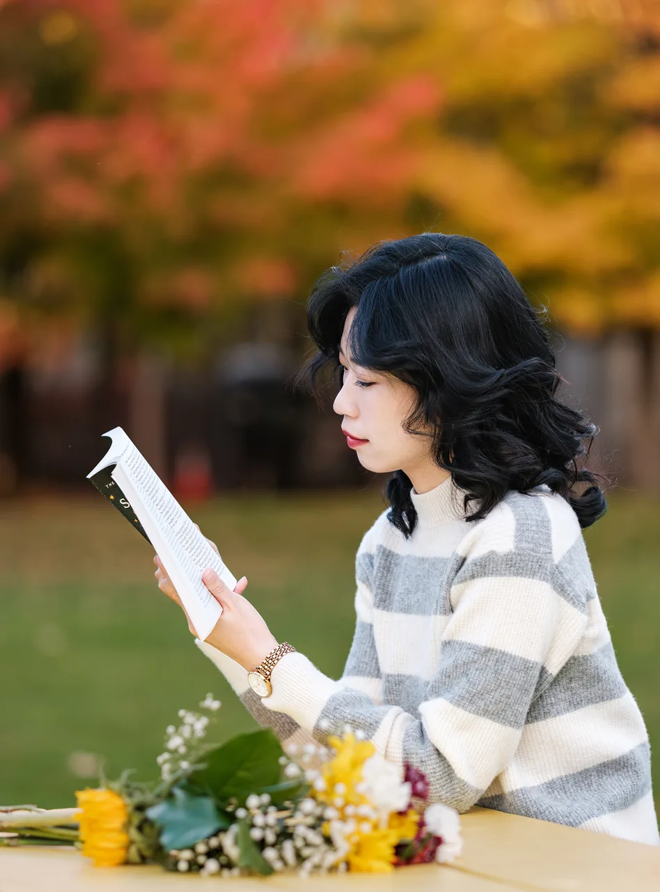 An outdoor portrait reading a book beside autumn foliage.